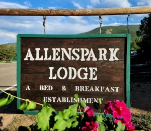 Sign for Allenspark Lodge, a rustic wooden sign with white lettering, surrounded by trees and mountains in the background.